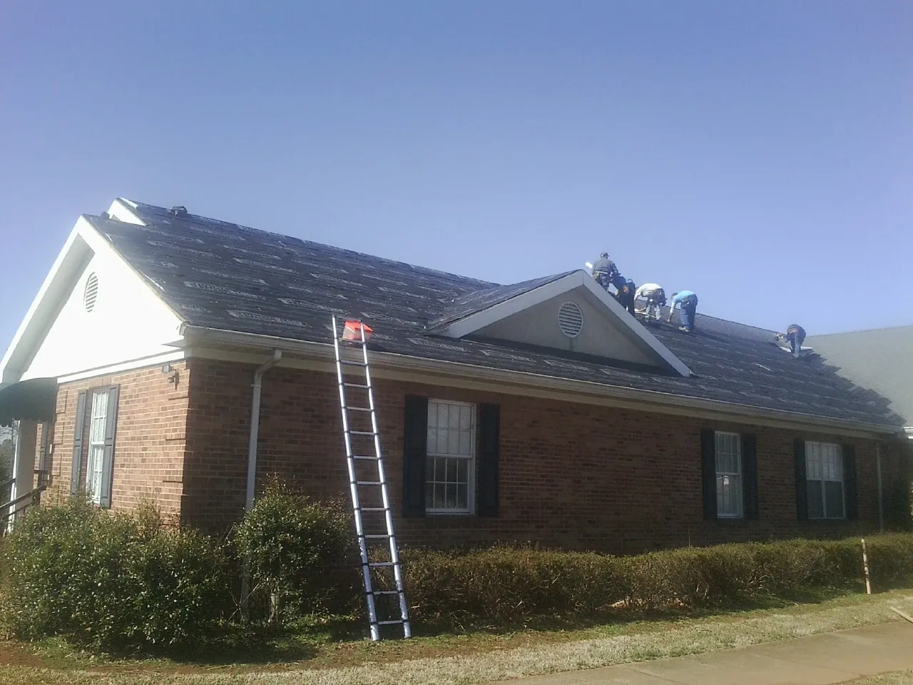 Crew tearing shingles off a brick ranch roof with a ladder set — Metal Roof Repair work by our crew in Marine City