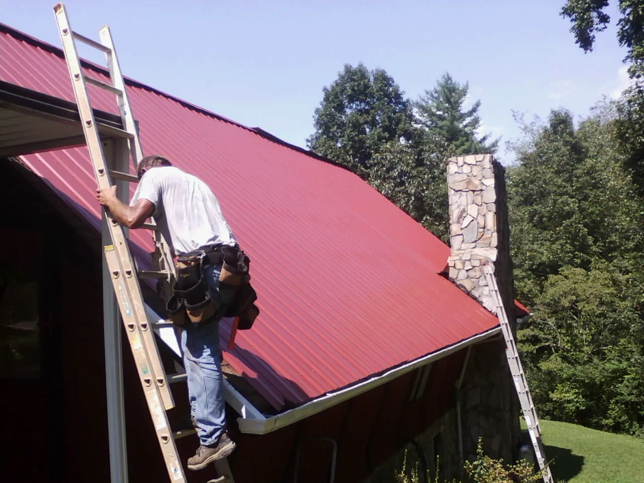 Roofer on a ladder beside a red metal roof and stone chimney — Soffit & Fascia Repair by Roofing Brothers in Marine City